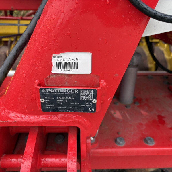 Red metal frame of agricultural equipment with a barcode label, a black Pöttinger plate, and hanging chains on the right side. Water droplets dot the surface, with yellow machinery parts faintly in the background.