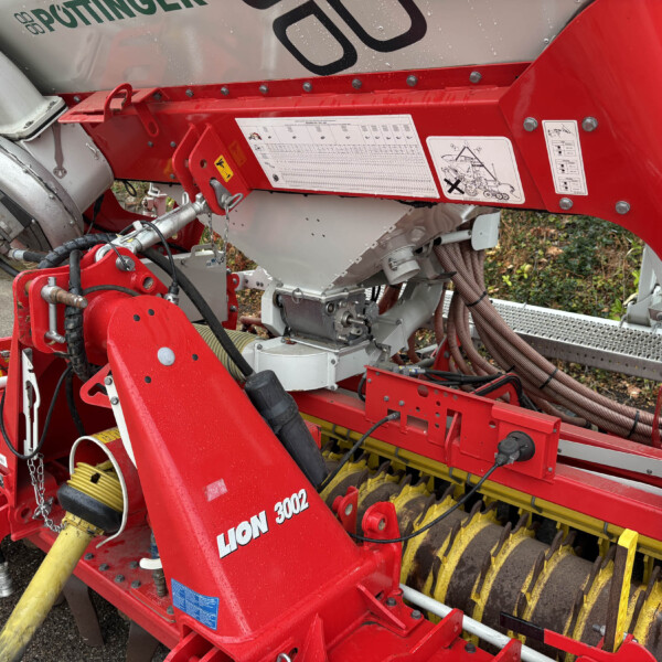 Close-up of a red agricultural machine with hydraulic hoses, warning labels, and a sticker reading 'LION 3002' on the side. Yellow rollers and metal components are visible in the foreground and background.