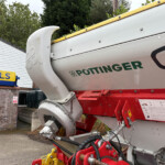 Close-up of a Pottinger agricultural machine with a silver body, red frame, and yellow rail, parked beside a brick building with a blue and yellow sign.