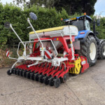 Blue tractor attached to a red-and-white seed drill with multiple discs, parked beside a hedge on a paved road.