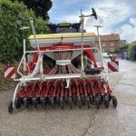 Rear view of a red and white seed drill attached to a tractor, with yellow top cover, hoses and tines, on a street beside a hedge and brick buildings.