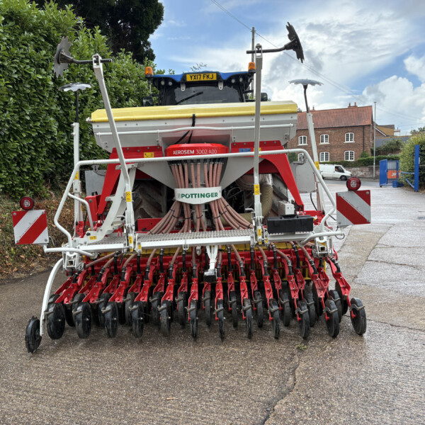 Rear view of a red and white seed drill attached to a tractor, with yellow top cover, hoses and tines, on a street beside a hedge and brick buildings.