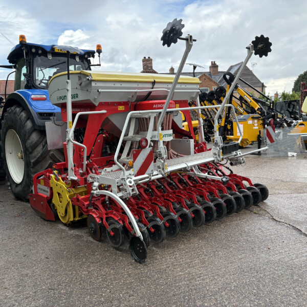 Blue tractor parked beside a long row of red and yellow farming implements in a yard, with houses in the background under a partly cloudy sky