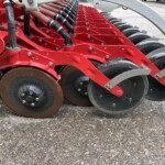 Close-up of a red farm implement with multiple metal discs and hydraulic components arranged in a row, ready for soil cultivation.