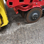 Close-up of a red construction machine’s frame with brown steel wheels beside a yellow attachment on an asphalt surface.