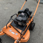 Orange commercial lawn mower deck with black engine and warning stickers mounted on a paved surface.