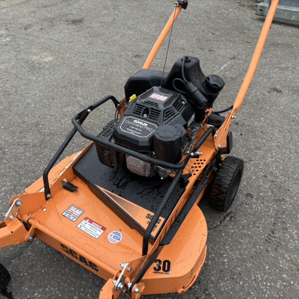 Orange commercial lawn mower deck with black engine and warning stickers mounted on a paved surface.