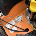 Close-up of an orange machinery panel with bolts, cables, and a wet surface; a yellow handle is visible nearby and labels are on the panel.