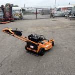 Orange SCAG lawn care machine in the foreground of a cluttered outdoor equipment yard with a metal fence and parked vans in the background