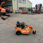 Orange turf cutter in the foreground with a green metal warehouse and parked equipment in the background.