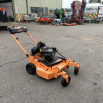 Orange SCAG lawn mower in a gravel lot, with industrial equipment and a white van behind a metal fence in the background.