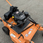 Orange outdoor machine with a black Kohler engine cover, yellow choke lever, and safety warning labels on a gray asphalt surface.