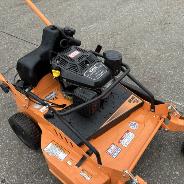 Orange outdoor machine with a black Kohler engine cover, yellow choke lever, and safety warning labels on a gray asphalt surface.