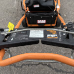 Top-down view of a yellow tag attached to a black and orange mower deck, showing control levers and safety warnings on the panel labeled SCAG Finish Cut.
