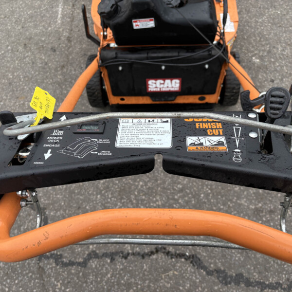 Top-down view of a yellow tag attached to a black and orange mower deck, showing control levers and safety warnings on the panel labeled SCAG Finish Cut.