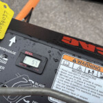 Close-up of a mower deck control panel with a yellow maintenance tag, an hours display reading 57, and orange warning labels on a wet surface.