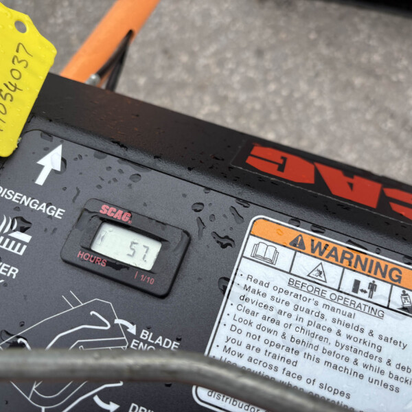Close-up of a mower deck control panel with a yellow maintenance tag, an hours display reading 57, and orange warning labels on a wet surface.