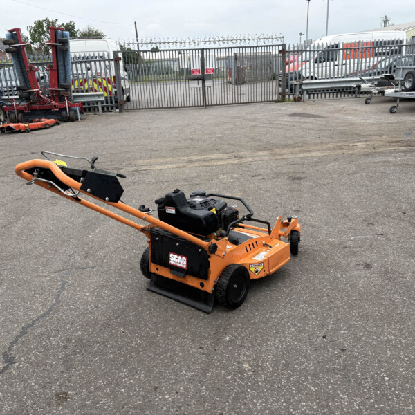 Orange SCAG lawn care machine in the foreground of a cluttered outdoor equipment yard with a metal fence and parked vans in the background