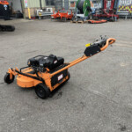 Orange maintenance machine in the foreground of an industrial yard, with various equipment, tires, and a fence in the background