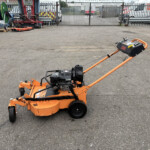 Orange lawn mower with a long handle in an industrial yard, with a metal fence, pallets, and equipment in the background