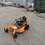 Orange SCAG stand-on mower in an outdoor industrial yard near a green dumpster and parked trailers in the background.