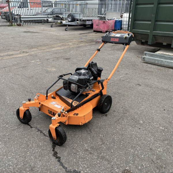 Orange SCAG stand-on mower in an outdoor industrial yard near a green dumpster and parked trailers in the background.