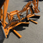 Orange industrial street-sweeper attachment with large black brush bristles on a paved surface, viewed up close.