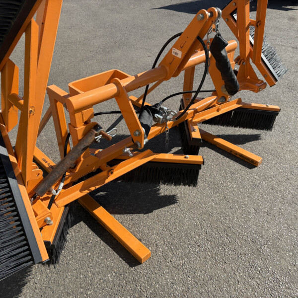 Orange industrial street-sweeper attachment with large black brush bristles on a paved surface, viewed up close.
