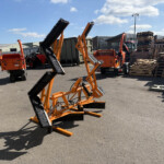 Industrial yard with orange road sweepers in the foreground, pallets stacked to the right, and a blue sky in the background.