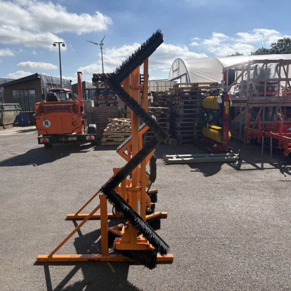 Orange industrial sweeper with a tall angled brush in the foreground, pallets and covered equipment in a yard behind it.