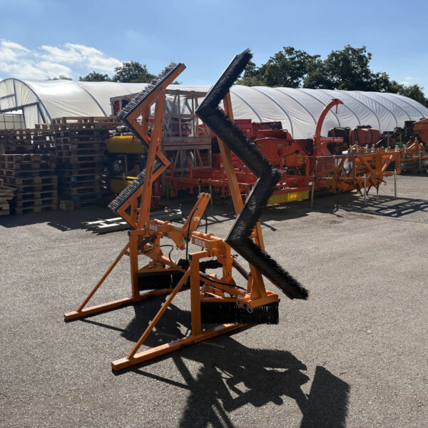 Orange agricultural machinery with large brush attachments in a yard, pallets stacked nearby and a white greenhouse in the background on a sunny day.