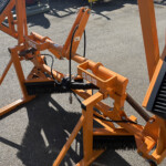 Orange industrial cleaning attachment with large cylindrical brushes and visible hydraulic hoses, resting on pavement.