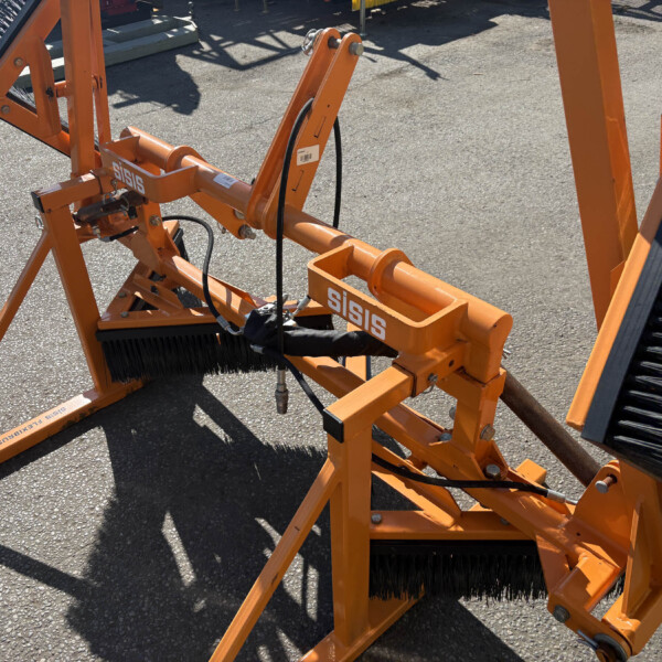 Orange industrial cleaning attachment with large cylindrical brushes and visible hydraulic hoses, resting on pavement.