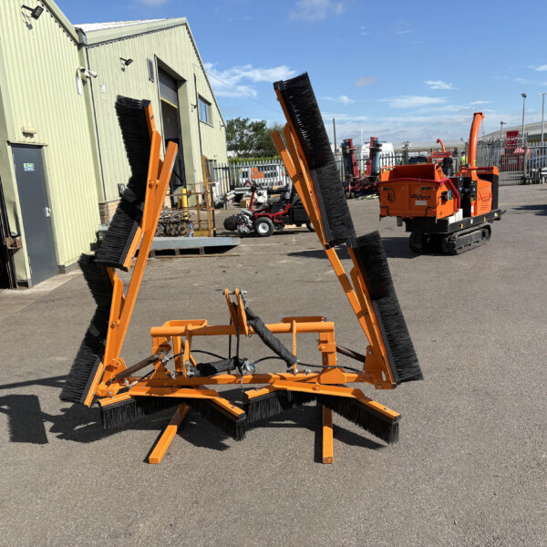 Orange industrial street-sweeper attachments in the foreground, with a pale green warehouse and clear blue sky in the background on an outdoor lot.