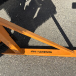 Close-up of an orange ISISIS FlexiBrush attachment on the ground, showing black bristles and stamped text along the beam.