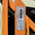 Close-up of a bright orange steel beam with a silver identification plate attached, in a warehouse/factory setting.