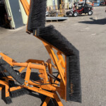 Orange industrial sweeping broom with dense black bristles mounted on a metal frame in an outdoor work yard.