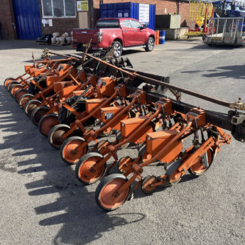 Line of orange farming equipment with wheels, arranged in a row in a parking lot, red pickup truck and industrial yard in background.