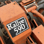 Close-up of an orange industrial machine part with bolts, showing dirt and wear and a black-and-white label partially reading text.