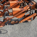 Close-up of orange agricultural machinery with multiple small wheels and rust on a gravel surface, likely a planter or cultivator frame.