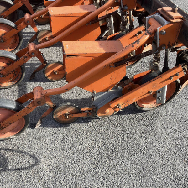 Close-up of orange agricultural machinery with multiple small wheels and rust on a gravel surface, likely a planter or cultivator frame.