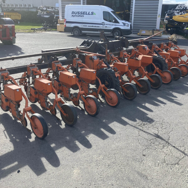 Row of orange farming implements with circular disks and metal frame parked outdoors, with a white van in the background.