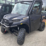Black off-road utility vehicle with large knobby tires and a heavy front bumper parked outside a glass-fronted building, front-left angle.