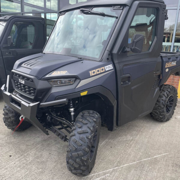Black off-road utility vehicle with large knobby tires and a heavy front bumper parked outside a glass-fronted building, front-left angle.