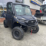 Dark blue off-road utility vehicle with large knobby tires parked outside a New Holland Agriculture dealership, signage visible on the building.