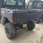 Rear of a dark off-road pickup with oversized mud tires and a tow hitch, license plate FX75 ZDS, parked beside another similar truck in a lot.