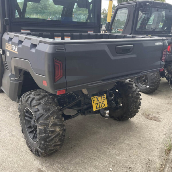Rear of a dark off-road pickup with oversized mud tires and a tow hitch, license plate FX75 ZDS, parked beside another similar truck in a lot.
