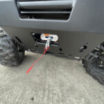 Front bumper of a rugged off-road vehicle with a winch and red tow strap attached, large tread tire visible on the left.