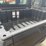 Dark gray pickup bed with a rugged plastic liner, stickers on the side, and a visible hitch ball; construction gear in the background sea of equipment.
