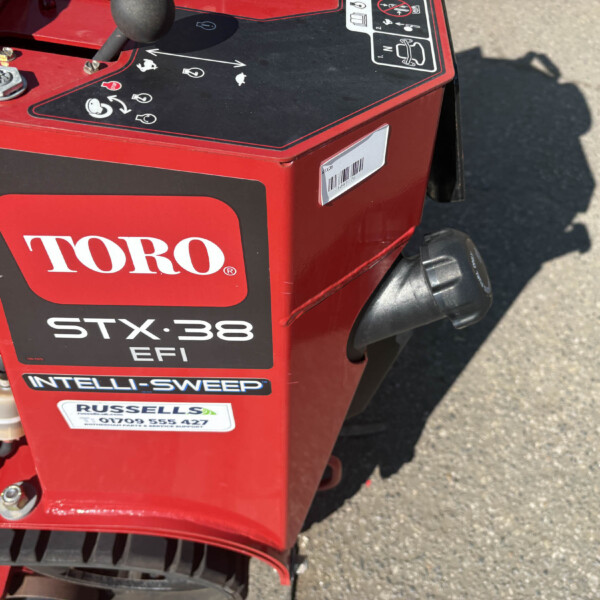 Close-up of a red Toro lawn tractor showing the TORO logo and STX38 EFI INTELLISWEEP branding on the side panel with a control panel and warning decals nearby.